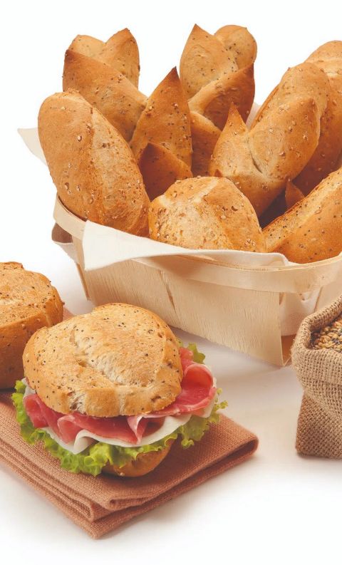 Multigrain bread rolls in a wooden basket, a filled sandwich with ham and lettuce, and a burlap sack of sesame seeds.