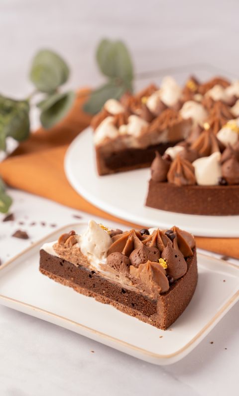 A slice of chocolate tart with piped cream and chocolate rosettes on a white plate, whole tart blurred behind on oran...