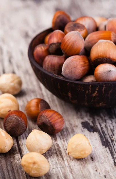 Whole hazelnuts in shells piled in a dark wooden bowl, with shelled and unshelled nuts scattered on a rustic wood sur...
