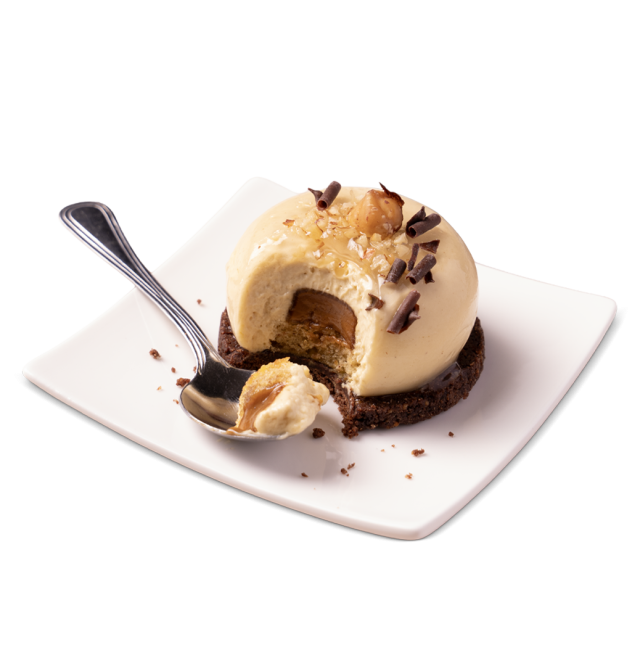 Domed caramel mousse dessert on a chocolate cookie base, topped with hazelnuts and chocolate curls, with a bite taken...