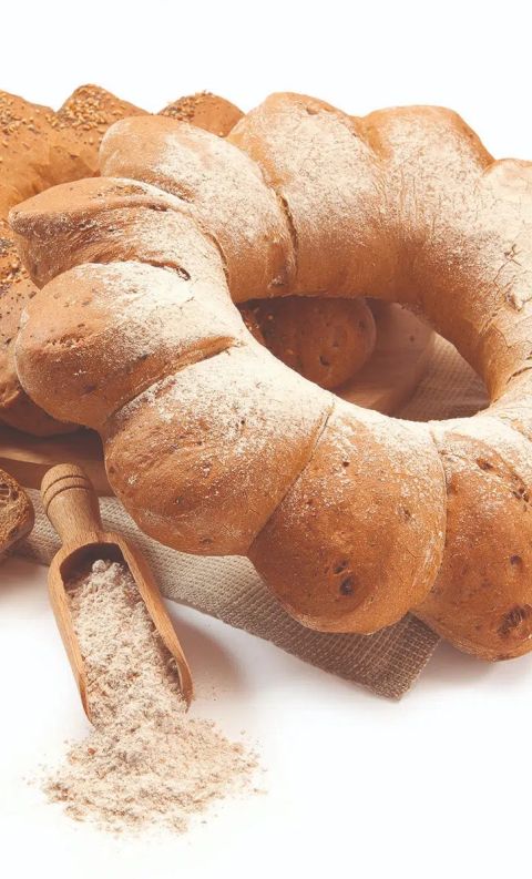A ring-shaped braided bread loaf dusted with flour, surrounded by seeded bread slices and a wooden scoop of flour.