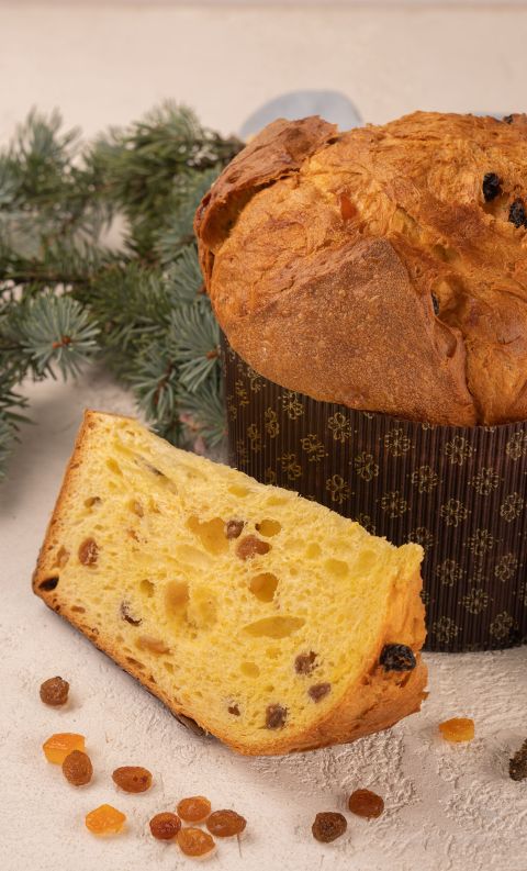 Panettone loaf in patterned wrapper with a sliced piece, raisins and candied fruit, pine branches behind.