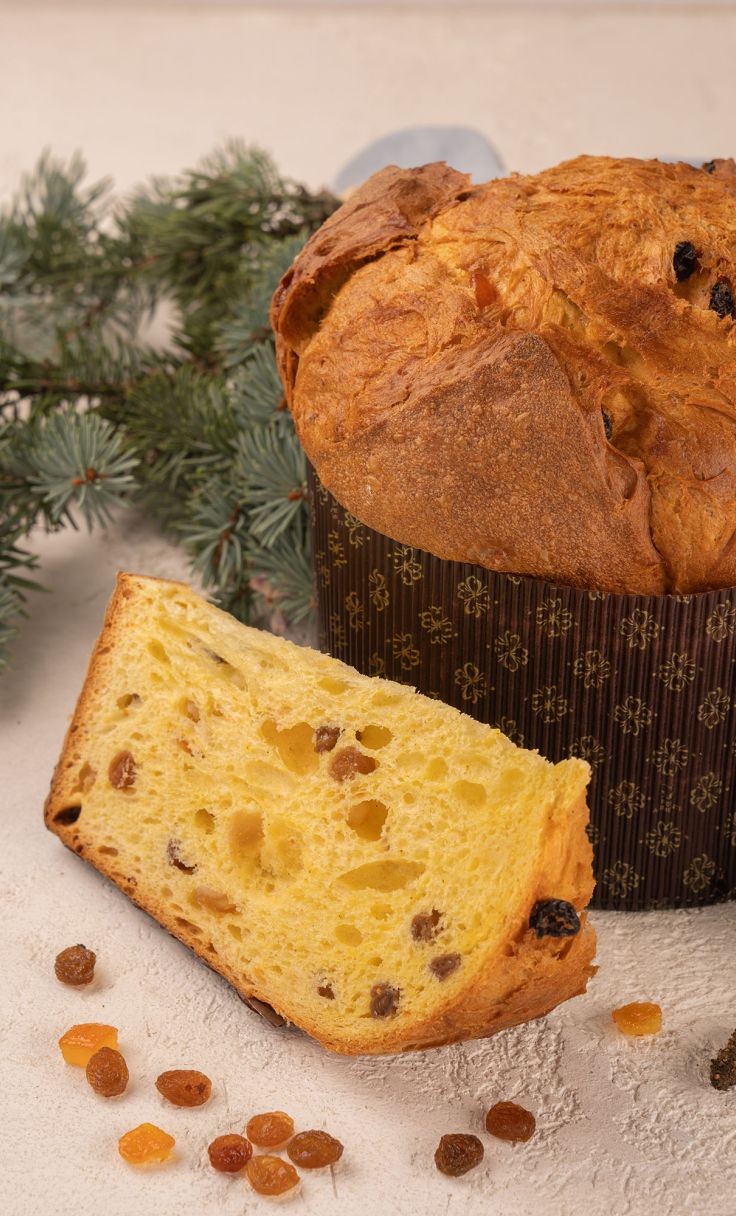 Panettone loaf in patterned wrapper with a sliced piece, raisins and candied fruit, pine branches behind.