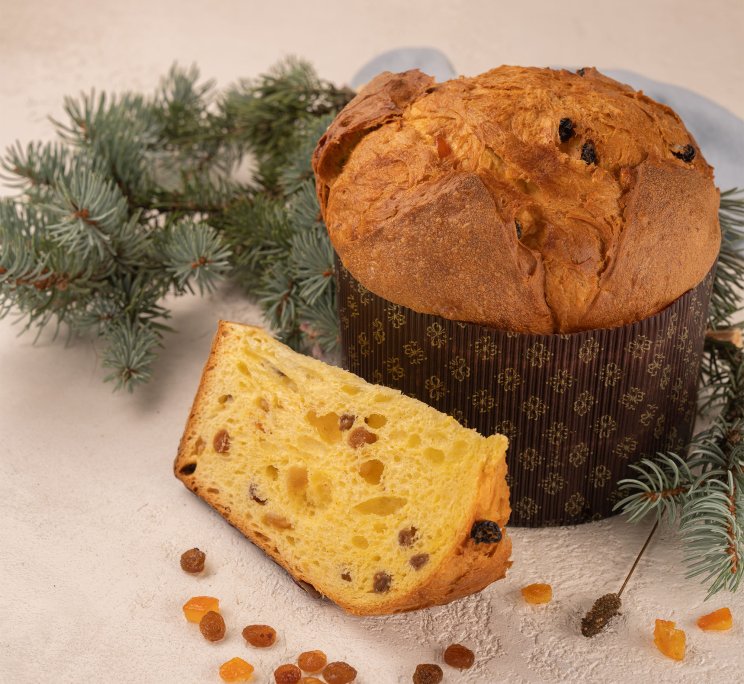 Panettone loaf in patterned wrapper with a sliced piece, raisins and candied fruit, pine branches behind.