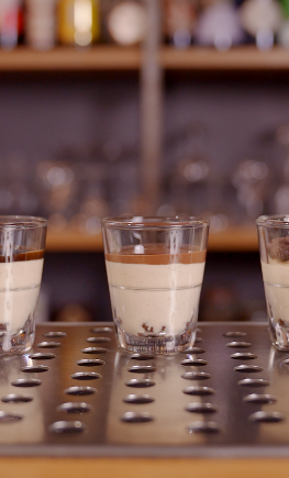 Three shot glasses of layered cream liqueur on a perforated bar counter, one topped with cocoa chunks