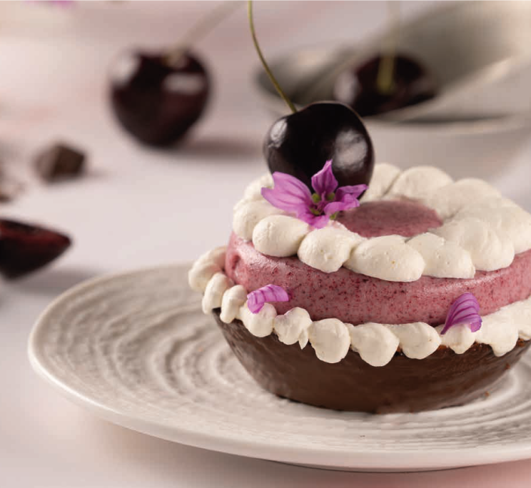 A chocolate tart with pink cherry mousse, piped cream, a fresh cherry, and purple flower petals on a white plate.