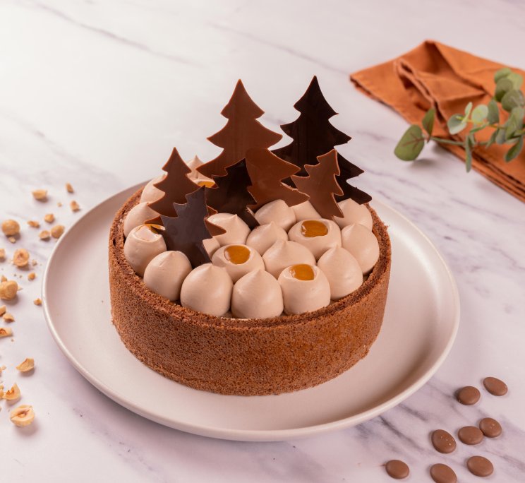 A round tart topped with cream swirls, caramel-filled dots, and chocolate tree decorations, surrounded by hazelnuts o...