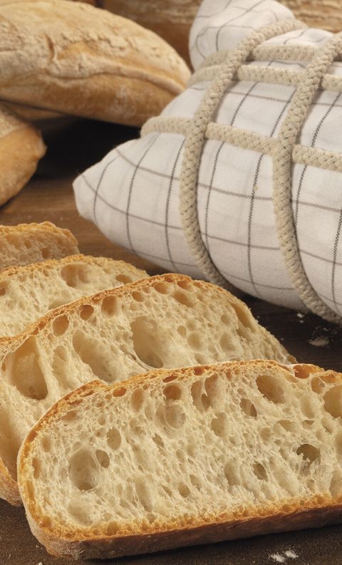 Sliced artisan bread loaves with an airy crumb, beside a checkered cloth-wrapped loaf tied with cord on a floured woo...