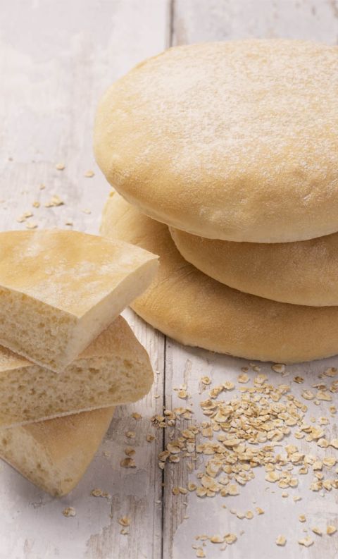 Stacked round flatbreads beside sliced pieces showing soft interior, with scattered oats on a white wooden surface.