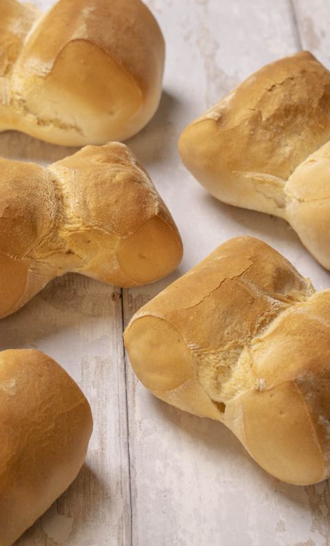 Five golden-brown bow-tie shaped bread rolls dusted with flour, arranged on a rustic white wooden table.