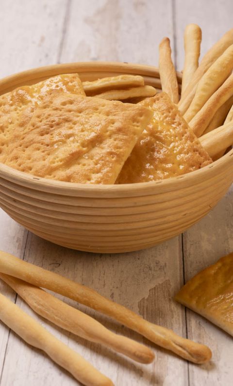 Wooden bowl with crispy flatbread crackers and breadsticks, surrounded by loose breadsticks on a light wooden table.