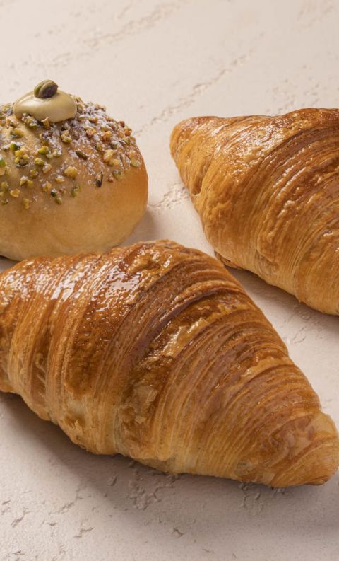 Two golden croissants beside a round pastry topped with crushed pistachios, cream, and powdered sugar.