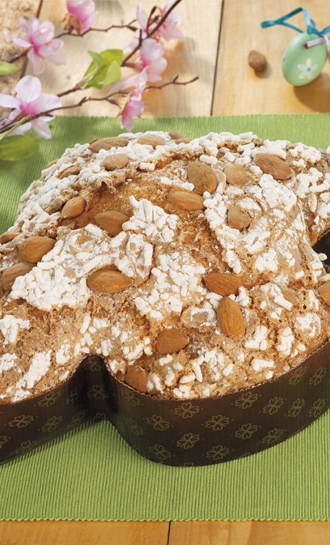 Dove-shaped Italian Easter cake (colomba) topped with almonds and sugar crystals, on a green mat with pink blossoms.