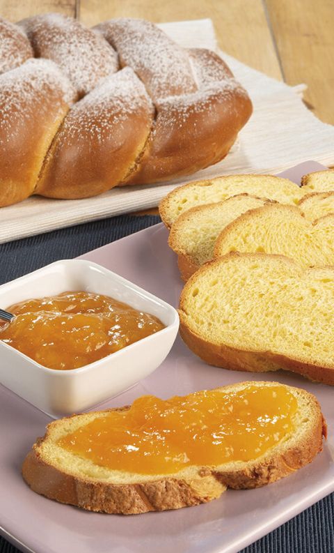 Braided sweet bread loaf with sliced pieces on a pink plate, one topped with apricot jam beside a small bowl of jam.