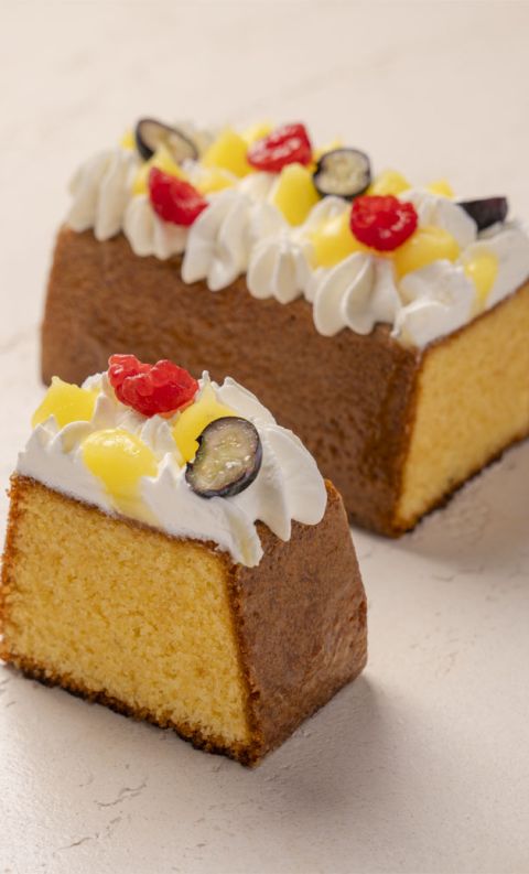 A whole loaf cake and a cut slice topped with whipped cream, candied pineapple, cherries, and dark grapes.