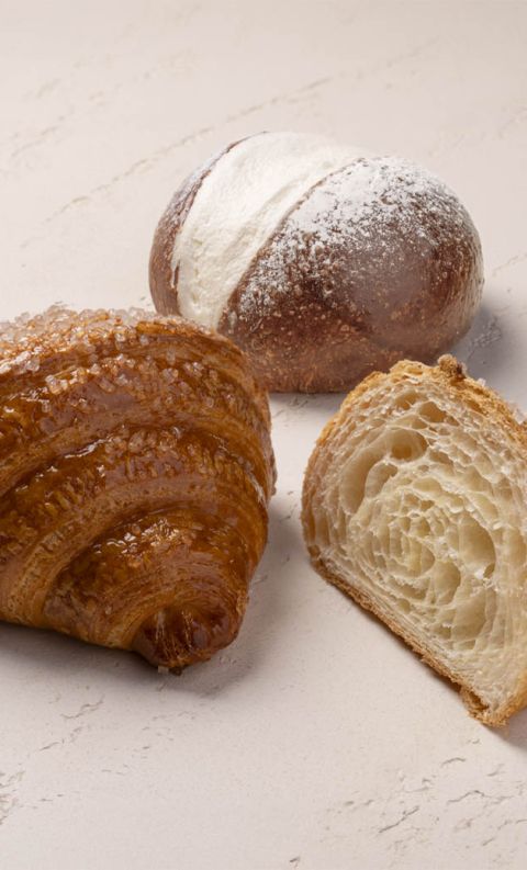A sugar-topped croissant sliced in half beside a powdered sugar-dusted pastry roll on a light stone surface.