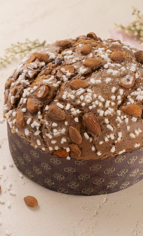 Round panettone topped with whole almonds and pearl sugar, in a dark patterned wrapper on a light surface with sprigs...