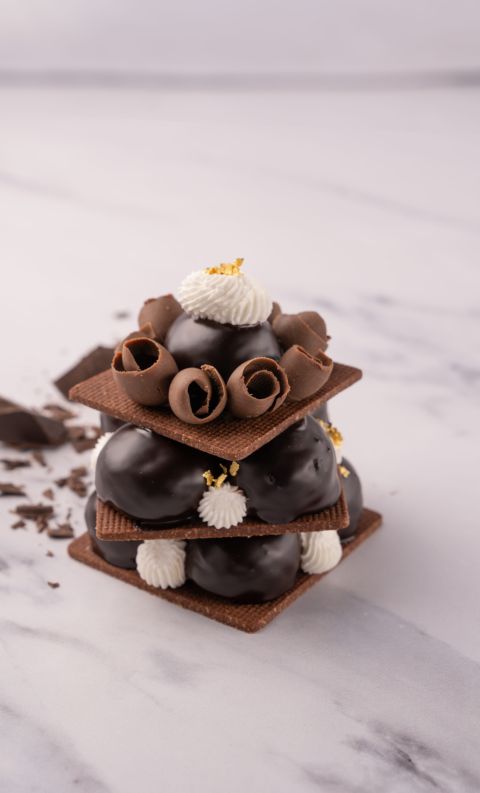 A layered chocolate pastry tower with cream puffs, chocolate curls, and thin wafer squares, garnished with gold leaf ...