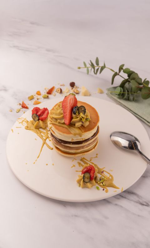 A stacked pancake dessert topped with piped caramel cream, sliced strawberries, and nuts, plated with a spoon and euc...