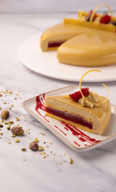 A slice of glossy yellow mirror-glazed cake with raspberry filling, garnished with cream and berries, beside pistachios.