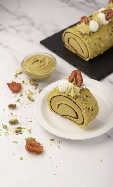 Pistachio cake roll slice with swirled filling on a white plate, garnished with cream, pistachios, and dried strawber...