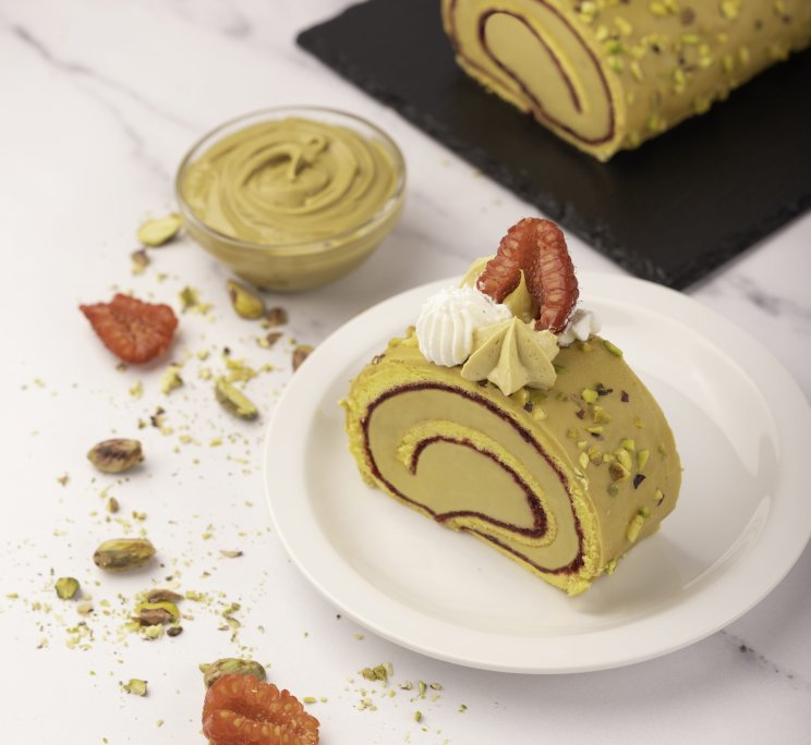 Pistachio cake roll slice with swirled filling on a white plate, garnished with cream, pistachios, and dried strawber...