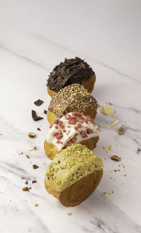 Four round pastries in a diagonal row, topped with dark chocolate shavings, chopped nuts, crushed freeze-dried berrie...