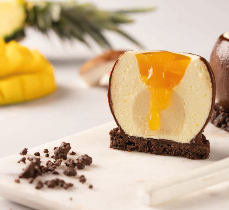 A cross-sectioned chocolate dome dessert with mango filling on a brownie base, garnished with cookie crumbs and fresh...