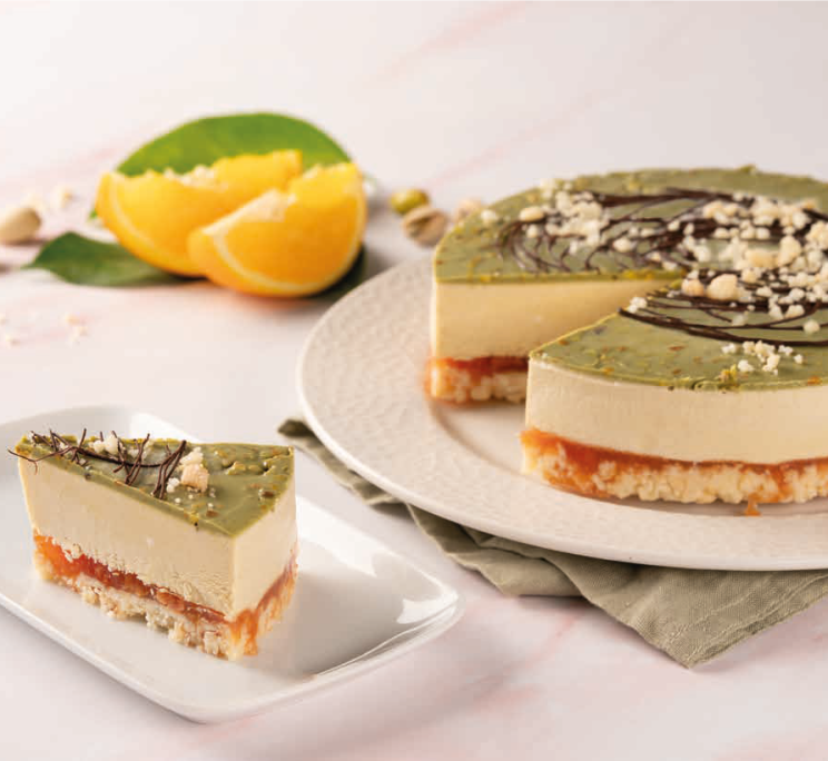 A layered cheesecake with pistachio top, cream filling, and orange glaze base, served whole and sliced beside orange ...