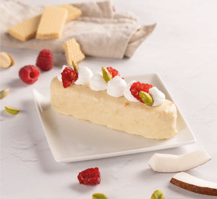 A wedge of pale cream cake topped with piped cream, raspberries, and pistachios, served with wafer cookies and coconu...