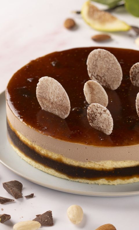 Round cake with chocolate mousse and glossy caramel glaze, topped with chocolate curls, on a plate surrounded by almo...