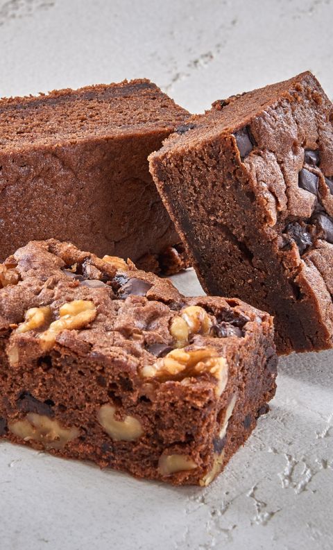 Three chocolate brownie squares with walnuts and chocolate chunks stacked on a light textured surface.