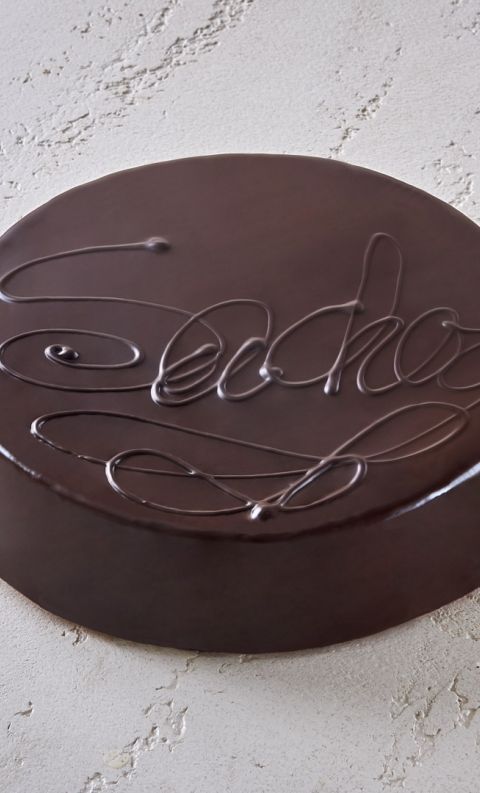 Round dark chocolate cake with glossy ganache glaze and decorative script piping on top, set on a textured white surf...