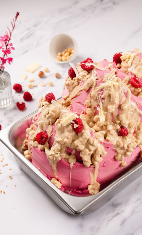 Pink frosted cake in a metal tray, topped with caramelized nut clusters, white chocolate shavings, and fresh raspberr...